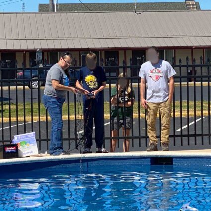 Four people are standing beside an outdoor pool, with some equipment on the pool's edge. Two persons are closely examining something held by the person in the middle. The background shows a building with a sign and parking spaces. Faces are blurred.