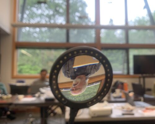 A camera capturing the reflection of a person in a small circular mirror. The person is inside a room with large windows revealing a green outdoor scenery. The room has tables with various items and appears to be a workspace or office.