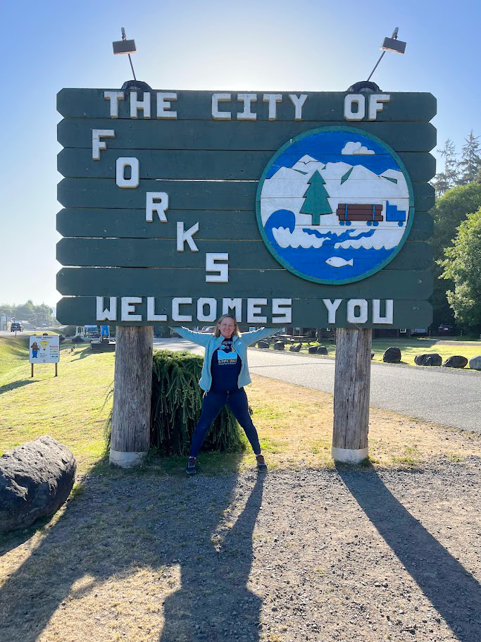 A person poses cheerfully with arms raised beneath a large wooden sign that reads "The City of Forks Welcomes You." The sign features a circular emblem depicting mountains, trees, and a river with logs. The surrounding area is green with trees and a paved road.