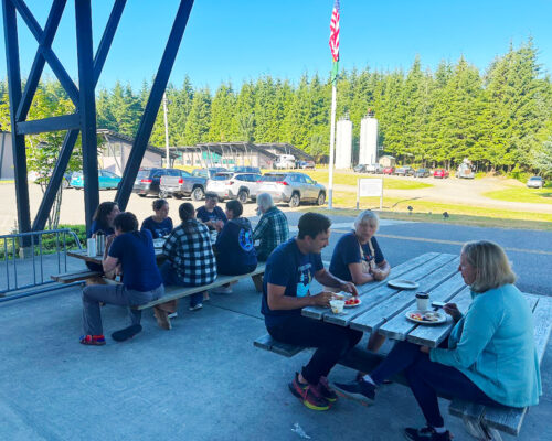 A group of people sits at wooden picnic tables outdoors, enjoying food and conversing. They are under a large pavilion near a parking lot with several cars and surrounded by green trees. The sky is clear, and an American flag is visible in the background.