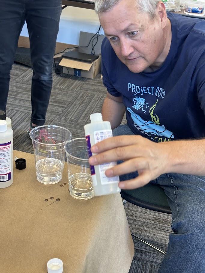 A person wearing a dark blue "Project R.O.C.K." T-shirt is carefully pouring a liquid from a bottle into a clear plastic cup. There are other bottles and clear cups on the table, which is covered with a brown paper sheet.