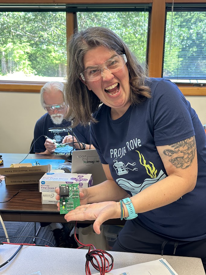 A woman wearing safety glasses smiling enthusiastically while holding a circuit board. She is in a room with large windows showing greenery outside. In the background, an older man with safety glasses and a beard is seen working on electronics.