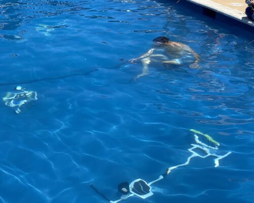 A person swimming underwater in a clear blue pool with submerged equipment nearby. The edge of the pool is visible in the background, and another person is partially visible above water near the edge.