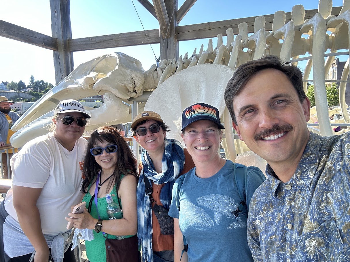 A group of five people smiling and posing in front of a large whale skeleton display at an outdoor location. They are dressed casually and appear to be enjoying a sunny day. Trees and other visitors are visible in the background.