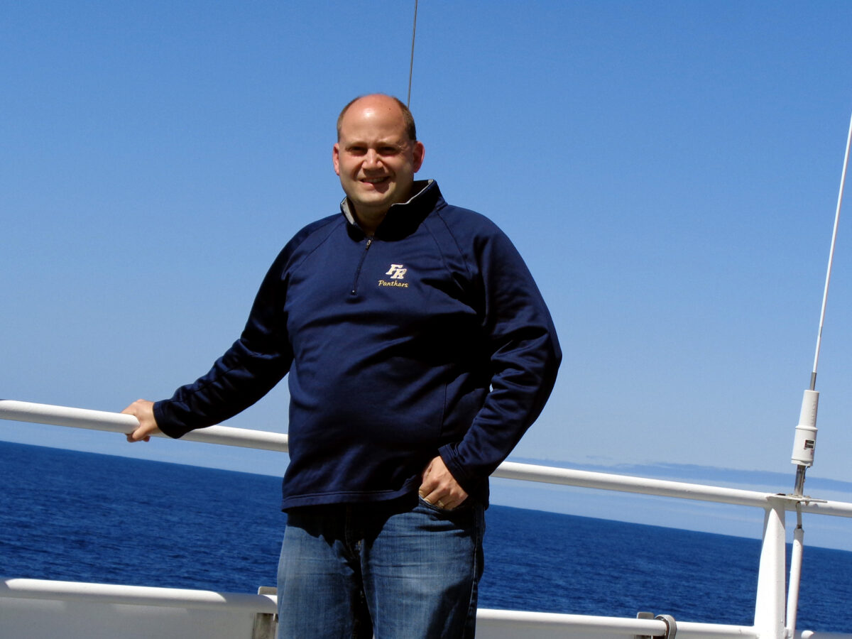 A person with a shaved head stands on a boat deck against a backdrop of the ocean and clear blue sky. They are wearing a dark blue sweatshirt and jeans, with one hand resting on the railing while the other is in their pocket.