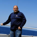 A person with a shaved head stands on a boat deck against a backdrop of the ocean and clear blue sky. They are wearing a dark blue sweatshirt and jeans, with one hand resting on the railing while the other is in their pocket.