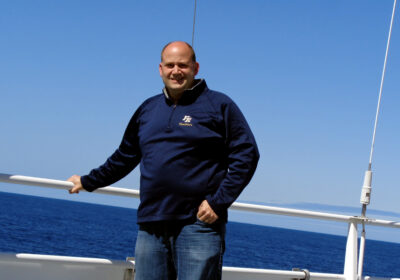 A person with a shaved head stands on a boat deck against a backdrop of the ocean and clear blue sky. They are wearing a dark blue sweatshirt and jeans, with one hand resting on the railing while the other is in their pocket.