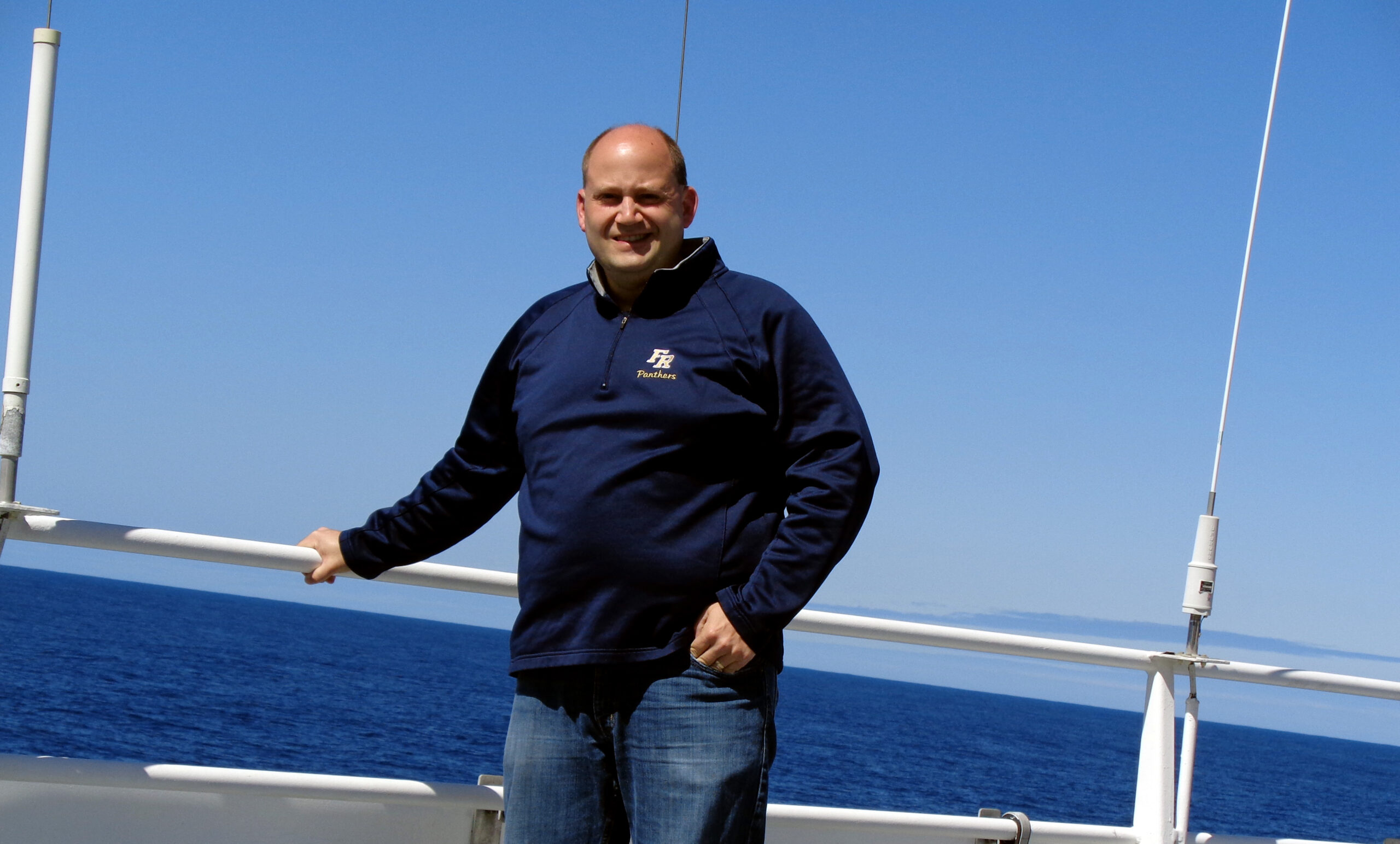 A person with a shaved head stands on a boat deck against a backdrop of the ocean and clear blue sky. They are wearing a dark blue sweatshirt and jeans, with one hand resting on the railing while the other is in their pocket.