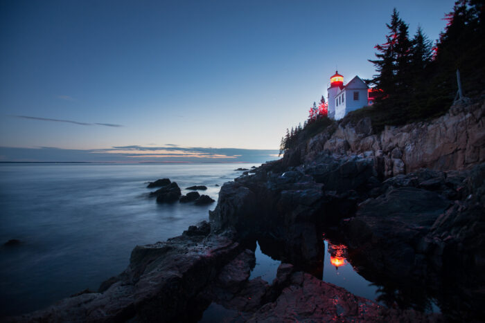 A serene coastal scene at dusk featuring a lighthouse perched on a rocky cliff, with its red beacon reflecting in the calm water below. The sky transitions from blue to the last light of sunset, and evergreen trees frame the lighthouse.