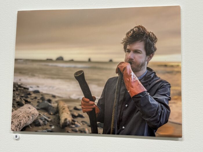 A man wearing an orange glove and dark overalls holds a tool by his side and gazes thoughtfully at the horizon. He stands on a rocky beach with driftwood and a calm ocean in the background, under a cloudy sky at sunset.