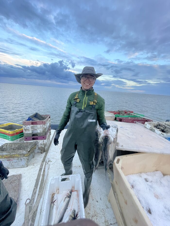 A person wearing green overalls, gloves, and a gray hat smiles while holding two large fish on a boat. The boat is surrounded by boxes and crates filled with fish. The background features a calm sea under a cloudy sky with hints of a sunset.