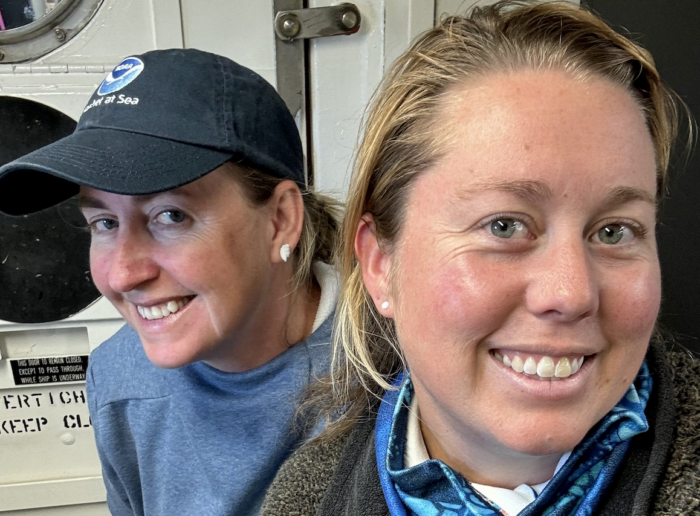 Two smiling people posing for a selfie. The person on the left is wearing a navy cap and a gray shirt. The person on the right is wearing a blue patterned neck gaiter and a brown hoodie. They are standing in front of a door with a circular window.