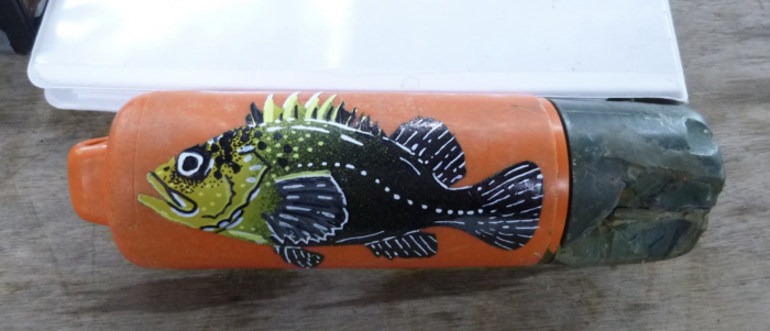 An orange and green cylindrical container with detailed fish artwork in black, white, and yellow. The illustrated fish has prominent fins and scales, and the container is resting on a wooden surface with a white paper or similar item partially visible behind it.