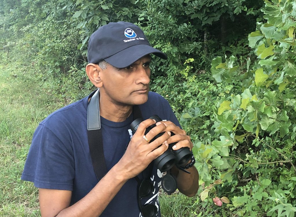 A person wearing a blue cap and shirt holds binoculars near dense green foliage, appearing focused. They have a black camera strap around their neck. The setting is outdoors with lush trees and plants in the background.