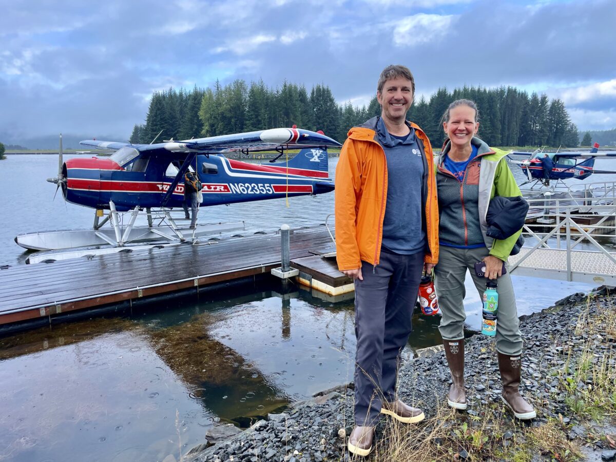 A man and woman stand smiling near a docked seaplane. They are dressed in outdoor gear, with the man wearing an orange jacket and the woman holding a water bottle. The background features another seaplane, a lake, and a forest under a cloudy sky.