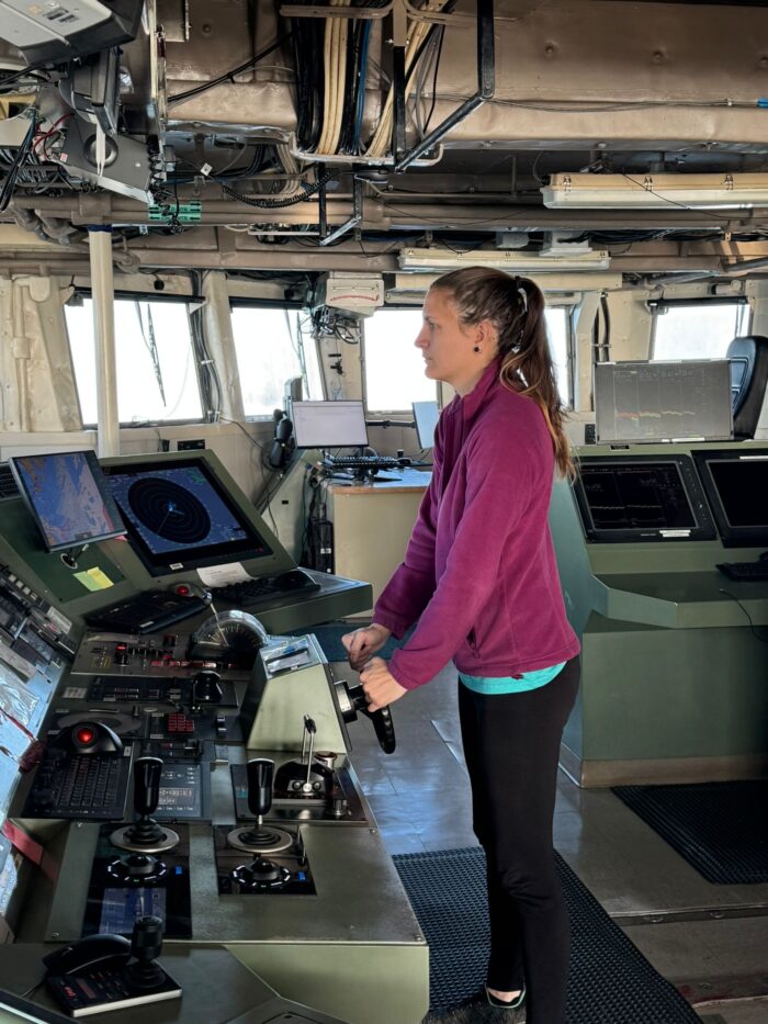 A person wearing a purple jacket operates controls on a ship's bridge, surrounded by navigation screens and equipment. They look forward, focused on steering or monitoring the vessel's systems.