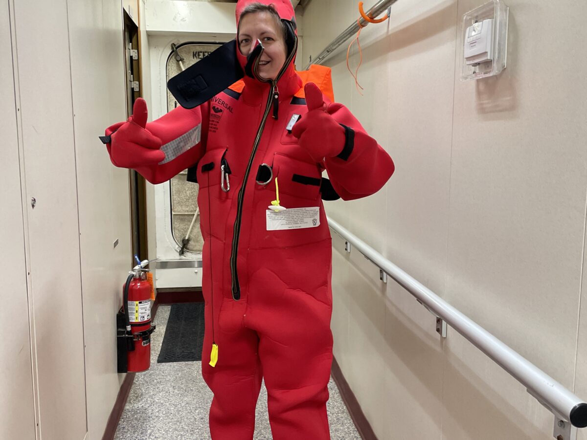 Person wearing an oversized red survival suit, giving a thumbs-up with both hands, stands in a narrow hallway of a ship. The suit is bright red with black zippers and patches. A fire extinguisher is mounted on the wall behind them.