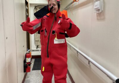 Person wearing an oversized red survival suit, giving a thumbs-up with both hands, stands in a narrow hallway of a ship. The suit is bright red with black zippers and patches. A fire extinguisher is mounted on the wall behind them.