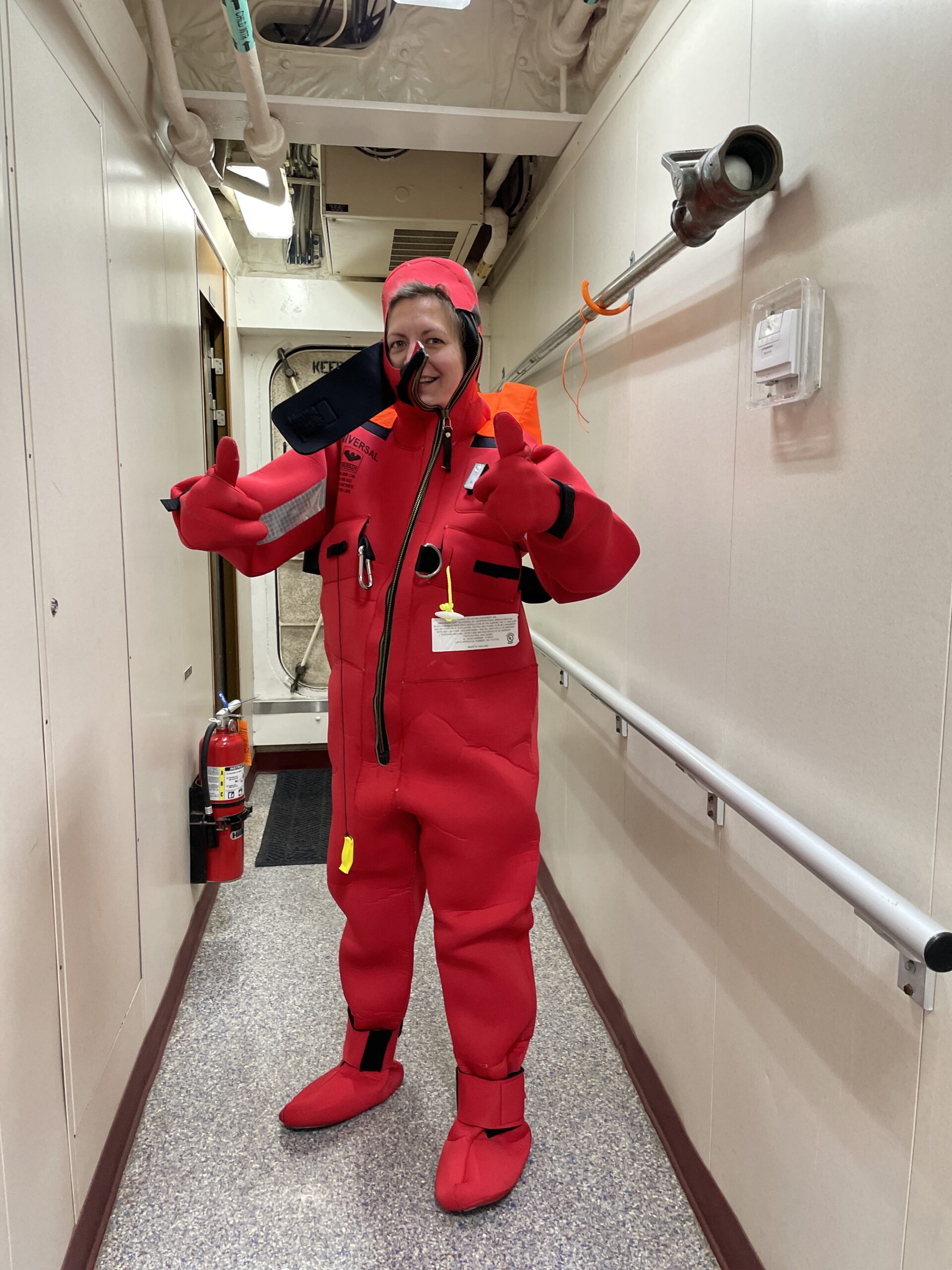 Person wearing an oversized red survival suit, giving a thumbs-up with both hands, stands in a narrow hallway of a ship. The suit is bright red with black zippers and patches. A fire extinguisher is mounted on the wall behind them.