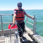 Person wearing a life jacket and sunglasses stands on a boat holding a metal frame. The water is calm and blue under a clear sky.