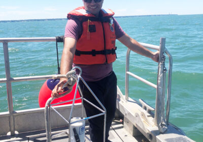 Person wearing a life jacket and sunglasses stands on a boat holding a metal frame. The water is calm and blue under a clear sky.
