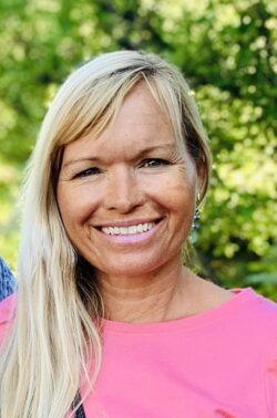 A person with long blonde hair is smiling warmly at the camera, wearing a pink top. The background is a blurred outdoor setting with green foliage.