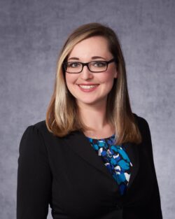A woman with shoulder-length blonde hair and glasses smiles at the camera. She is wearing a black blazer over a blue and white patterned blouse. The background is a plain, textured gray.