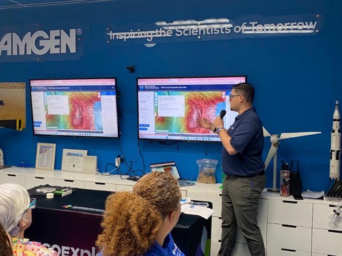 A man stands and speaks into a microphone while pointing at a screen displaying a topographic map. Several people sit and watch. The room has science posters, a wind turbine model, and a partial rocket model.