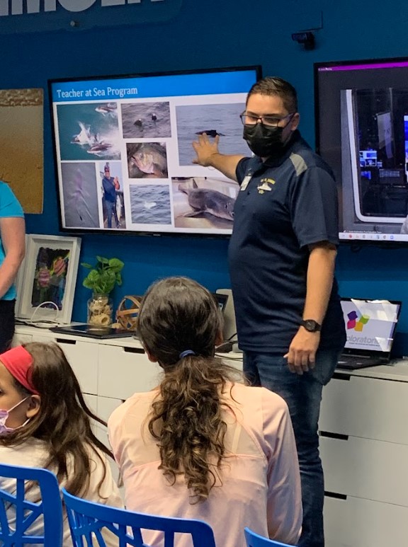 A masked man points to marine animal photos on a screen labeled "Teacher at Sea Program" while speaking to a group of children seated and watching in a classroom.