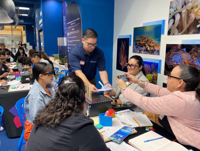 A man hands a booklet to a group of women seated at a table covered with books and science materials in a classroom decorated with ocean life posters. The women are engaged and interacting with each other.