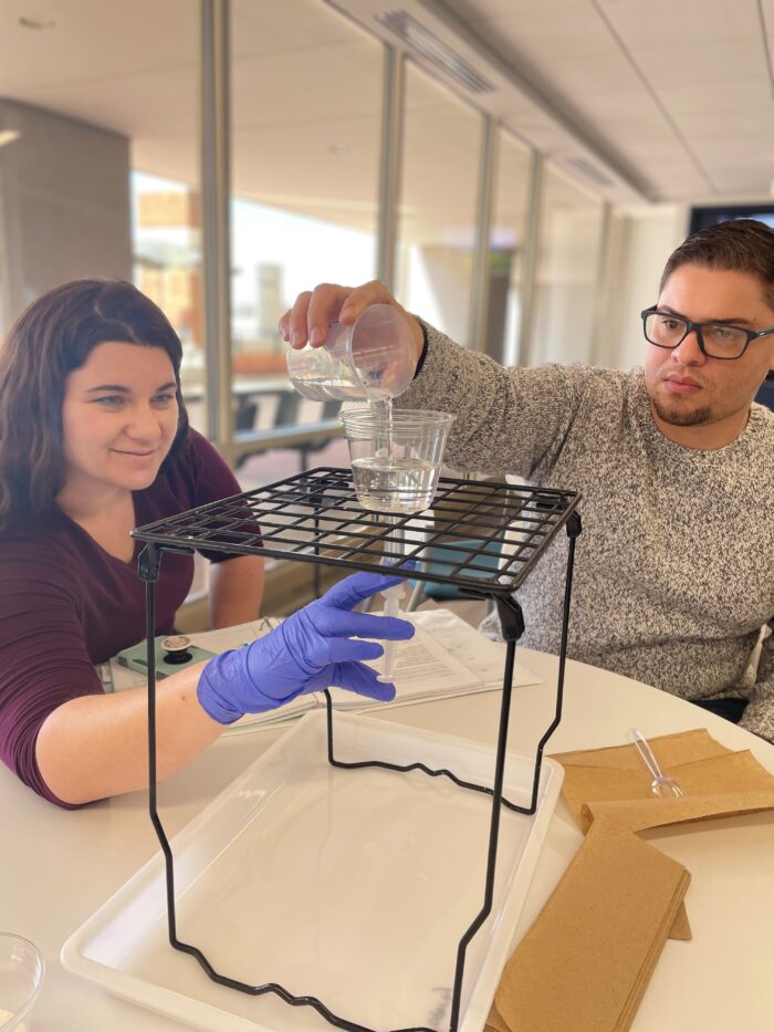 Two people conduct a science experiment indoors. One pours liquid from a beaker into a cup on a metal stand while the other, wearing a blue glove, adjusts the setup. Papers and utensils are on the table.