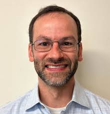 Smiling man with glasses, short dark hair, and a beard, wearing a light plaid shirt, stands against a plain beige background.