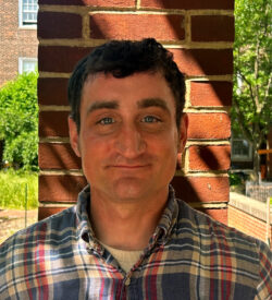 A person with short dark hair, wearing a checkered shirt, stands against a brick wall. The sunlight casts shadows on the wall, and greenery is visible in the blurry background.
