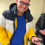 A smiling person wearing glasses and a yellow, white, and black jacket holds a small brown flat fish in their hand indoors.