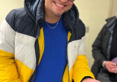 A smiling person wearing glasses and a yellow, white, and black jacket holds a small brown flat fish in their hand indoors.