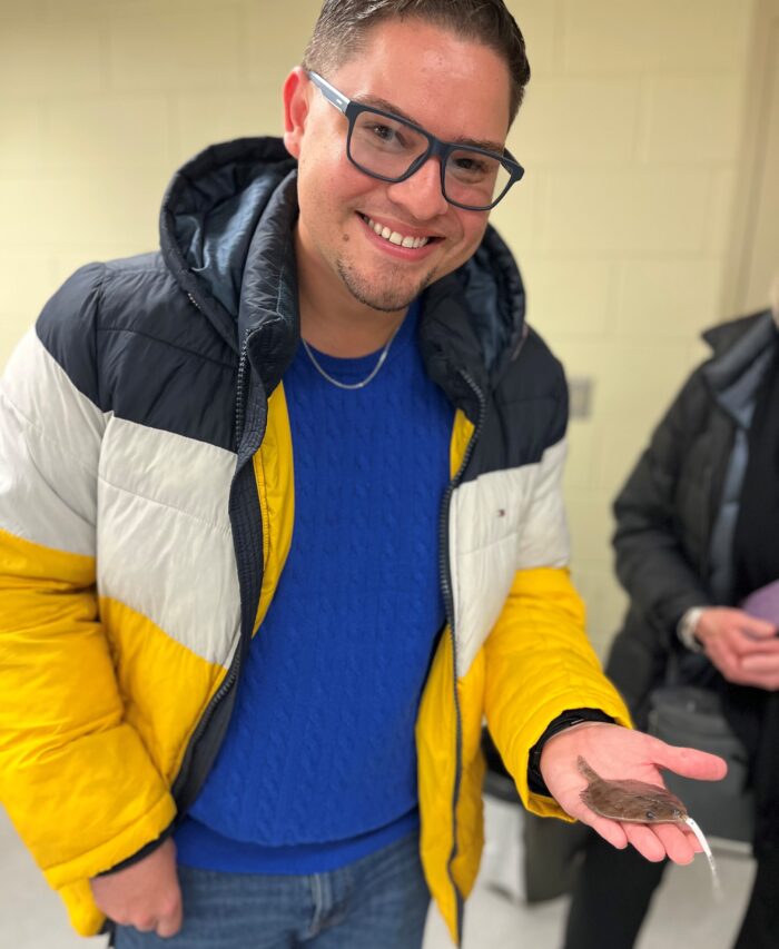 A smiling person wearing glasses and a yellow, white, and black jacket holds a small brown flat fish in their hand indoors.