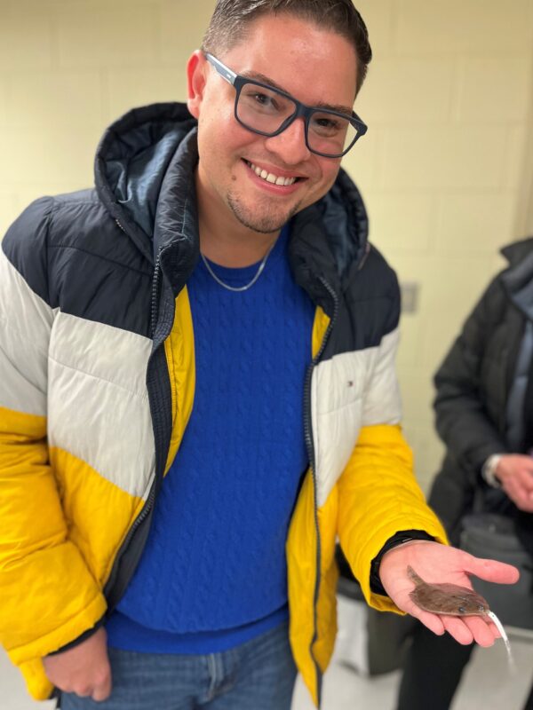 Michael-CR A smiling person wearing glasses and a yellow, white, and black jacket holds a small brown flat fish in their hand indoors.