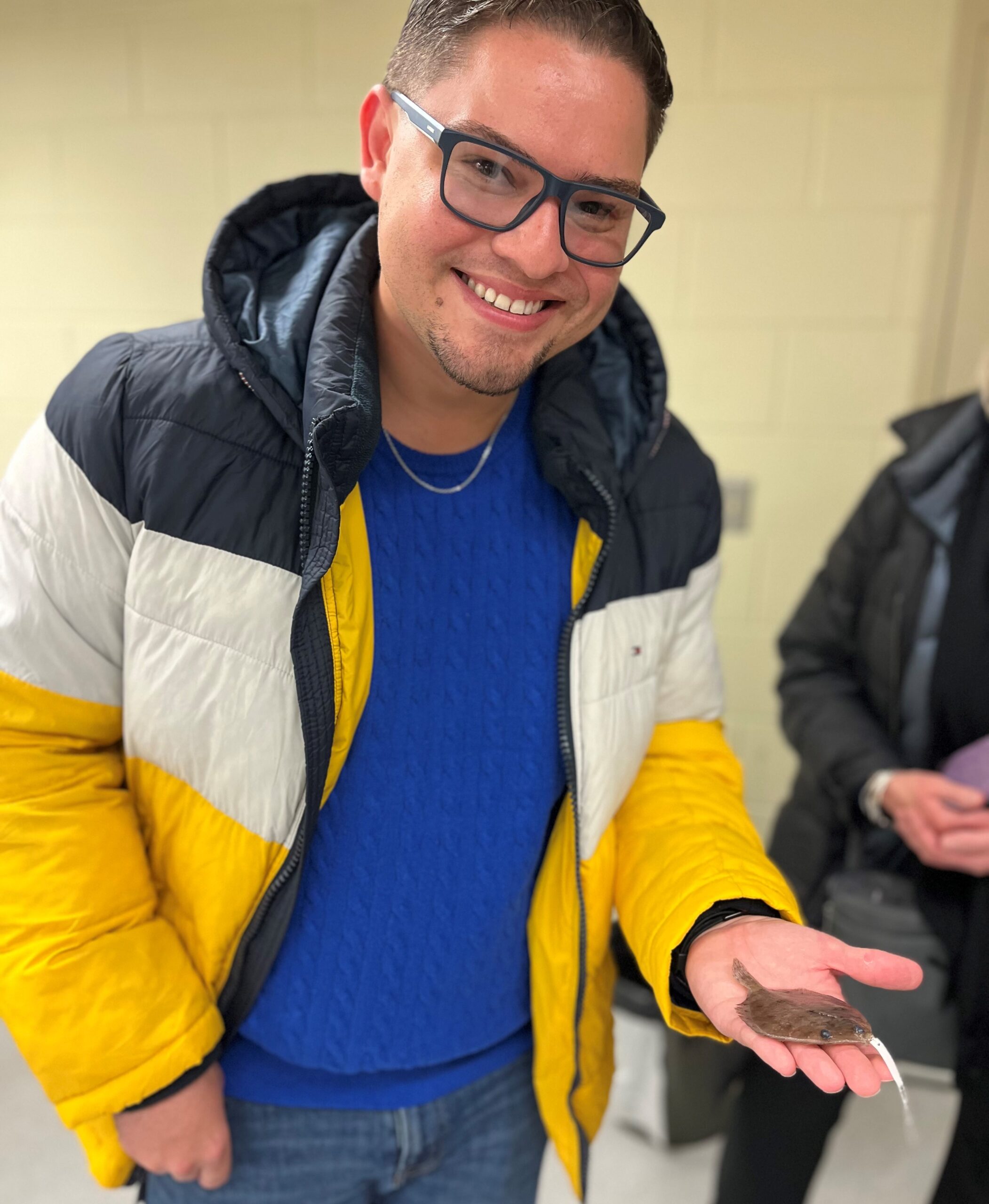 A smiling person wearing glasses and a yellow, white, and black jacket holds a small brown flat fish in their hand indoors.
