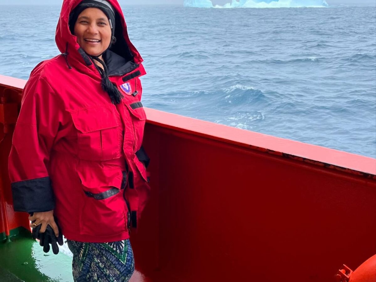 A woman in a red hooded jacket smiles while standing on the deck of a ship at sea, with an iceberg visible in the distance under a cloudy sky.