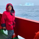 A woman in a red hooded jacket smiles while standing on the deck of a ship at sea, with an iceberg visible in the distance under a cloudy sky.