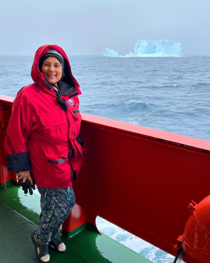 A woman in a red hooded jacket smiles while standing on the deck of a ship at sea, with an iceberg visible in the distance under a cloudy sky.
