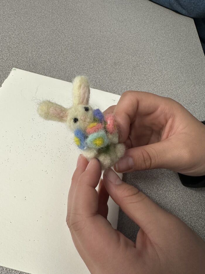 A person holds a small, handmade felted bunny with colorful decorations on its body, above a light gray surface next to a blank sheet of white paper.