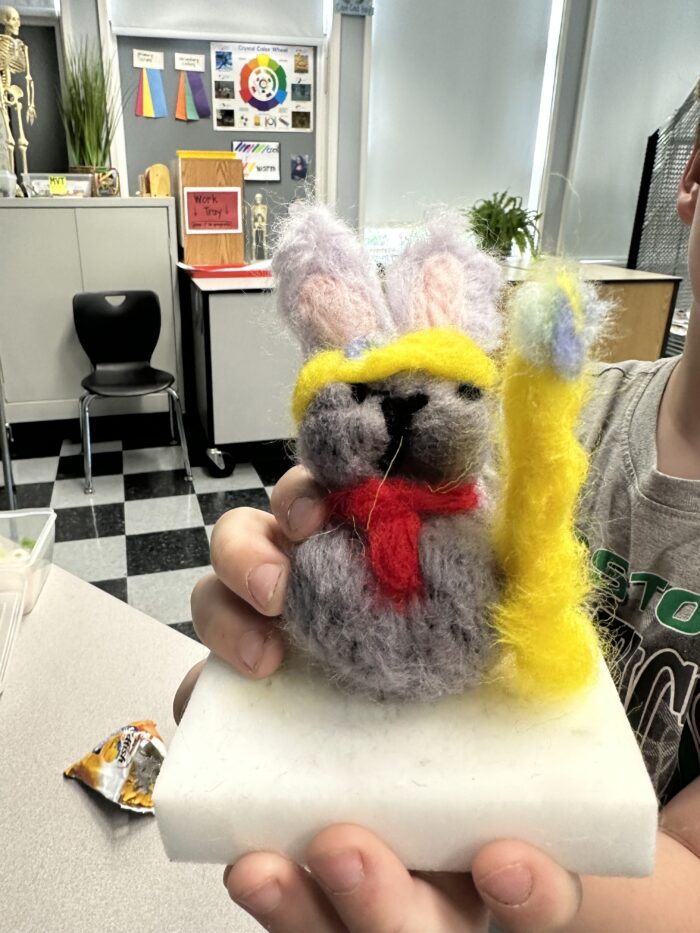 A hand holds a gray felted bunny with a yellow eye mask and yellow sword, displayed on a white foam base. The background shows a classroom with chairs, posters, and educational materials.