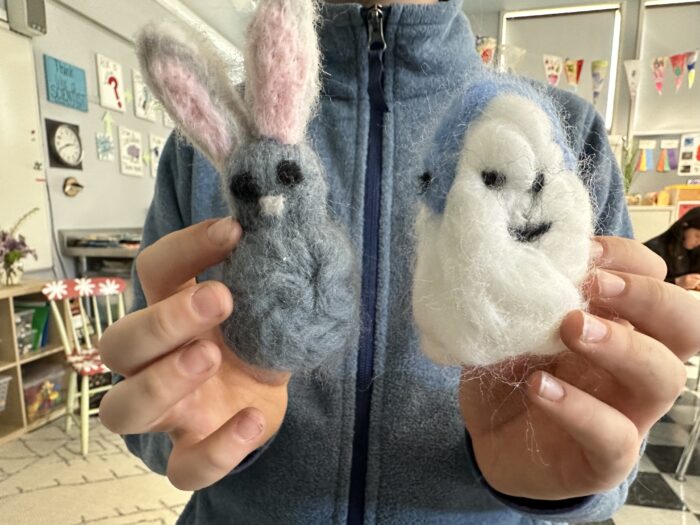 A person in a blue fleece holds two needle-felted wool figures: a gray bunny with pink ears and a white ghost with black eyes and mouth. Bright classroom decorations and art are visible in the background.