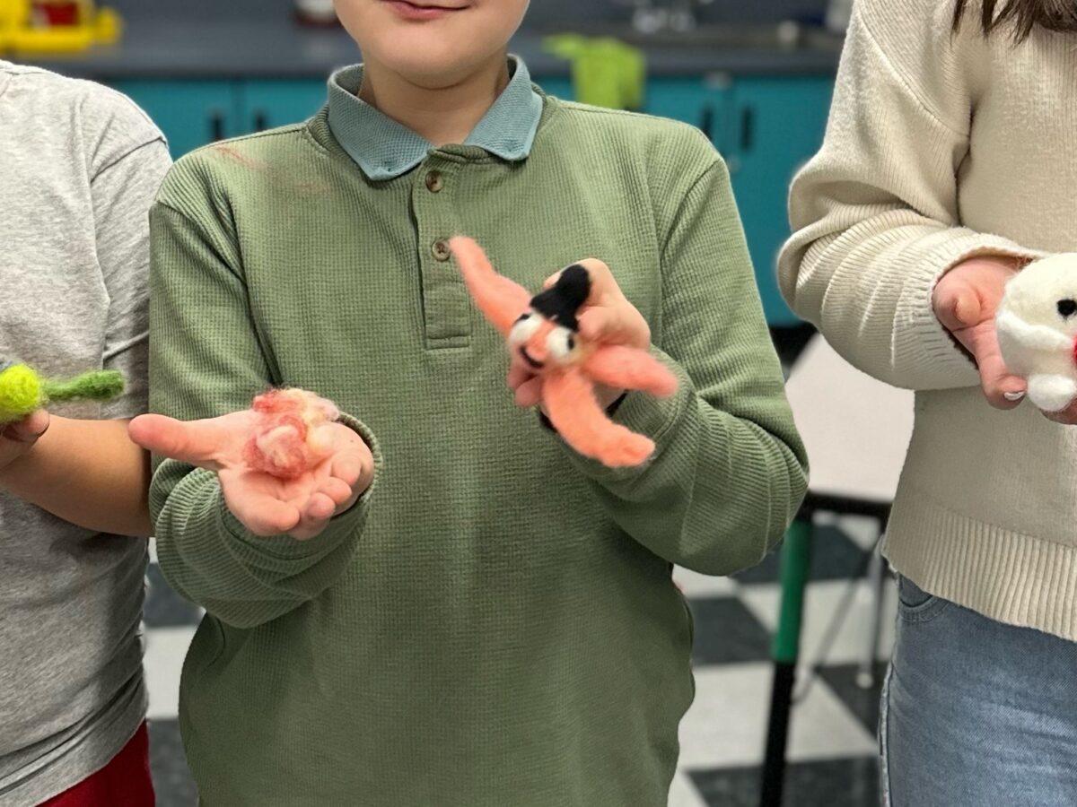 Three children stand side by side and hold up handmade felted animal figures in their hands, smiling and displaying the colorful, small creations in a classroom setting.
