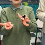 Three children stand side by side and hold up handmade felted animal figures in their hands, smiling and displaying the colorful, small creations in a classroom setting.