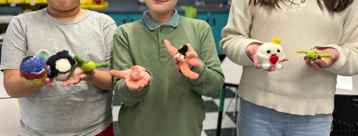 Three children stand side by side and hold up handmade felted animal figures in their hands, smiling and displaying the colorful, small creations in a classroom setting.