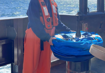 A person in orange waterproof overalls, a white hard hat, and a life vest stands on a boat deck by the ocean, holding a coiled blue rope on top of a metal post. The sky is clear and sunny.