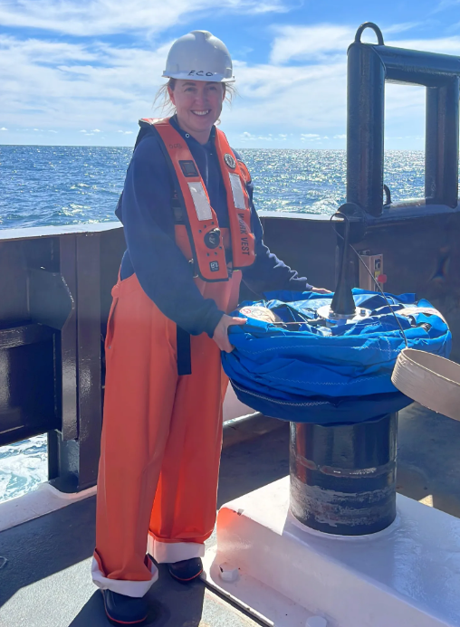 A person in orange waterproof overalls, a white hard hat, and a life vest stands on a boat deck by the ocean, holding a coiled blue rope on top of a metal post. The sky is clear and sunny.