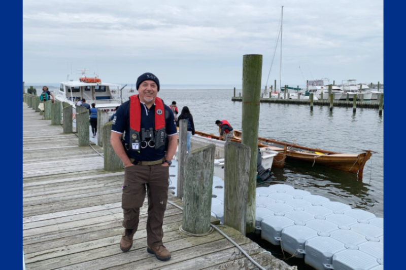 A smiling person in a life vest and beanie stands on a wooden dock by the water, with boats and people behind them on a cloudy day.